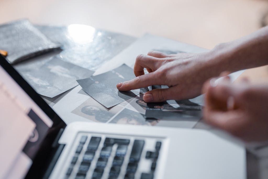 Prognose 2026 Ahnenforschung; Die Ahnenforschung wird digitaler. A hand arranges vintage photographs near a laptop in a modern workspace.