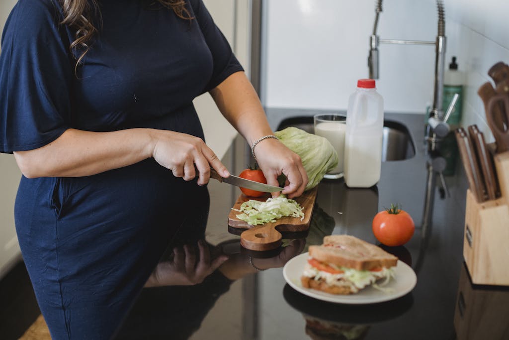 Crop anonymous pregnant female in blue dress standing near counter and chopping fresh lettuce leaves for sandwich in modern kitchen